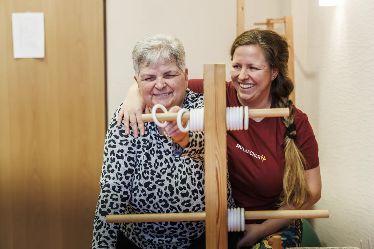 Therapeutin und ältere Patientin lachen gemeinsam bei einer Übung am Trainingsgerät