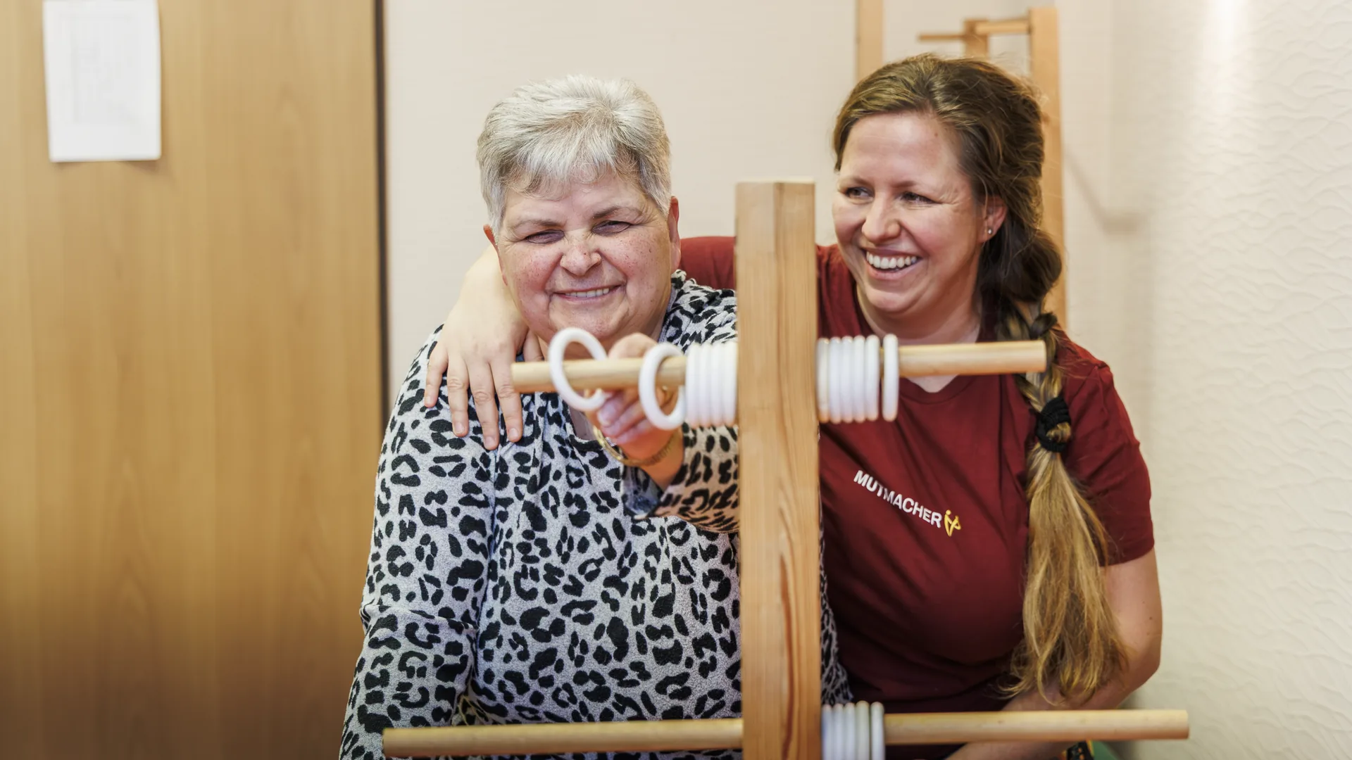 Therapeutin und ältere Patientin lachen gemeinsam bei einer Übung am Trainingsgerät