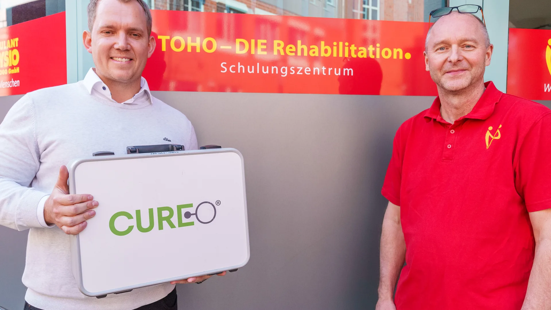 Zwei Personen mit einem Gerätekoffer vor dem Banner des TOHO-Schulungszentrums